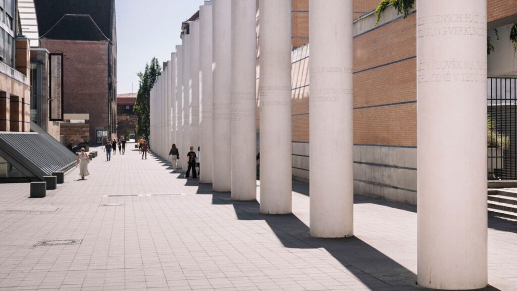Die Straße der Menschenrechte. Menschen laufen an den Säulen vorbei, die so hoch wie die Gebäude daneben sind