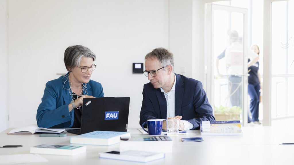 Katrin Kinzelbach und Markus Krajewski schauen gemeinsam in einen Laptop. Auf dem Tisch liegen Bücher und Papiere verstreut