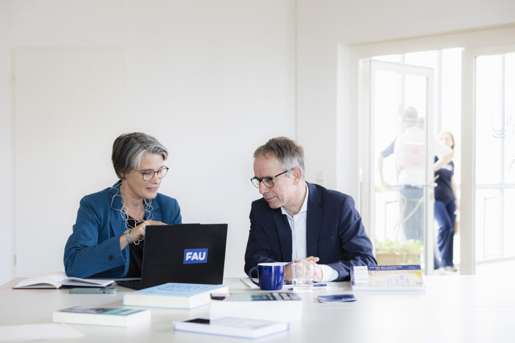 Katrin Kinzelbach und Markus Krajewski schauen gemeinsam in einen Laptop. Auf dem Tisch liegen Bücher und Papiere verstreut